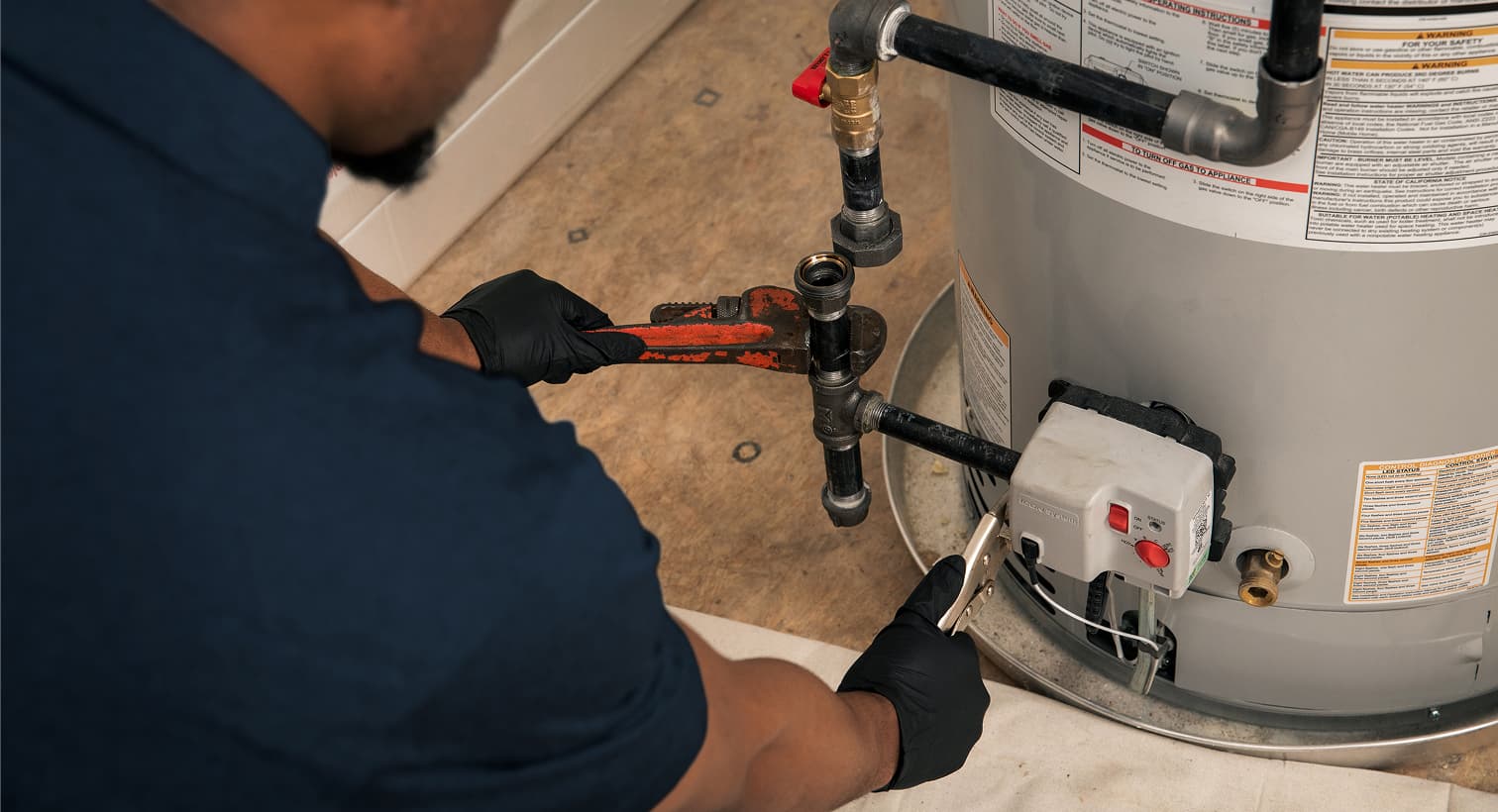 A Performance plumber wearing black gloves uses a pipe wrench to repair the gas line and control valve assembly on a residential water heater tank.