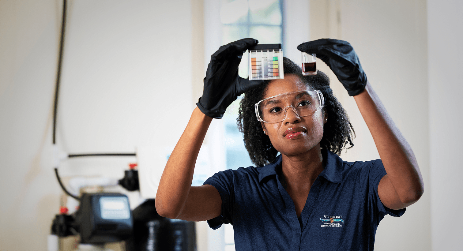 A Performance technician in a blue uniform and safety gear compares a water sample to a testing chart in front of a water softener system.